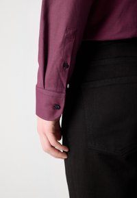 Personne portant une chemise bordeaux à manches longues avec des poignets boutonnés et un pantalon noir, main détendue le long du corps.