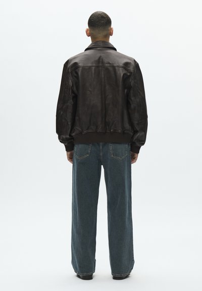 Férfi áll háttal, fekete bőrdzsekit és laza kék farmert visel, egyszínű világos háttér előtt.