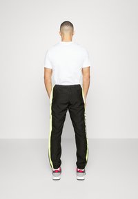 Mann med kort hår, iført T-skjorte i hvitt, svarte joggebukser med neongule sidestriper og hvite joggesko, stående vendt bort.