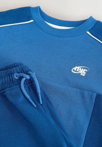 Sweat bleu avec logo blanc "nyc 82" sur la poitrine et pantalon de jogging bleu assorti avec cordon et taille élastique, présenté à plat.