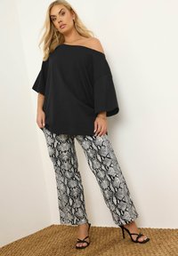 Svart off-shoulder topp i kombination med vita och svarta byxor med snakeskin-mönster och svarta sandaler med remmar, på en texturerad naturlig matta.