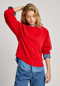 Felpa rossa con un fit rilassato, collo a costine e spalle scese, indossata sopra una camicia di jeans azzurro chiaro. Tessuto morbido con dettagli minimi.