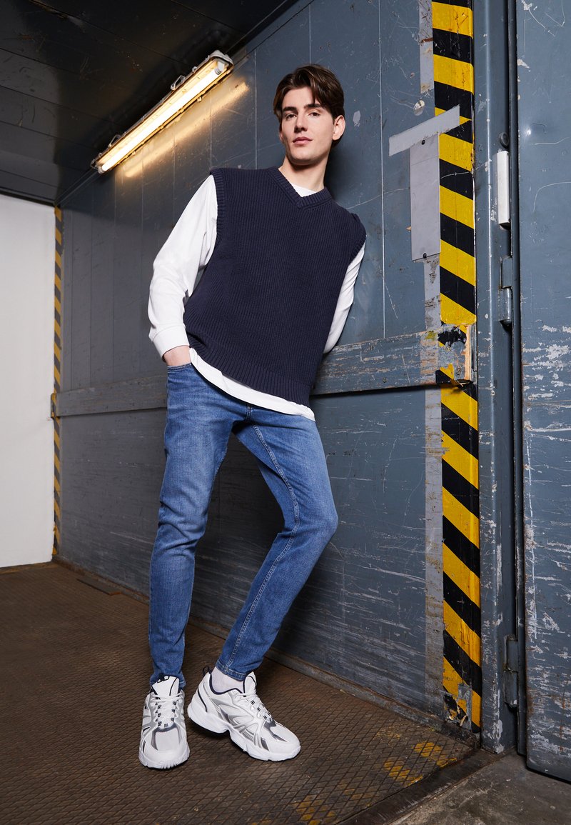 Chaleco de punto navy sobre una camisa blanca de manga larga, combinado con jeans azul claro y zapatillas de running blancas, con un telón de fondo industrial.
