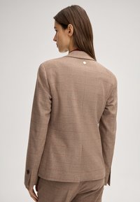 Blazer a quadri marrone con sottili linee rosse, spalle strutturate e un dettaglio di un singolo bottone metallico. Tessuto texturizzato con una vestibilità su misura.