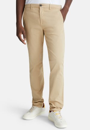 Beige chinos made of cotton blend, featuring slim fit, front pockets, a button closure, and paired with white sneakers.
