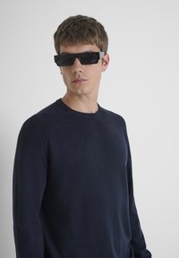 Zwarte rechthoekige zonnebril met grijze accenten, gedragen door een model in een donkerblauwe trui met getextureerde ribbel schouders, tegen een eenvoudige achtergrond.