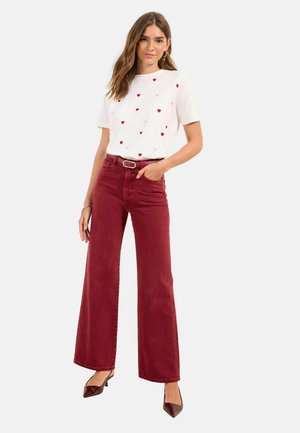 Kvinde iført hvid kortærmet skjorte med små røde hjerter, højtaljede, vidde bukser i rødt og brune spidse hæle, stående med hånden i lommen.