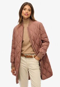 Femme portant une veste bomber matelassée mauve sur un pull à col roulé beige, associée à un pantalon beige clair et une ceinture tressée marron.
