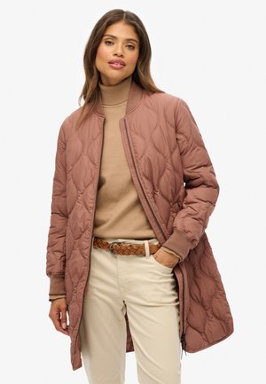 Vrouw in een gewatteerde mauve bomberjack over een beige coltrui, gecombineerd met lichtbeige broek en een gevlochten bruine riem.