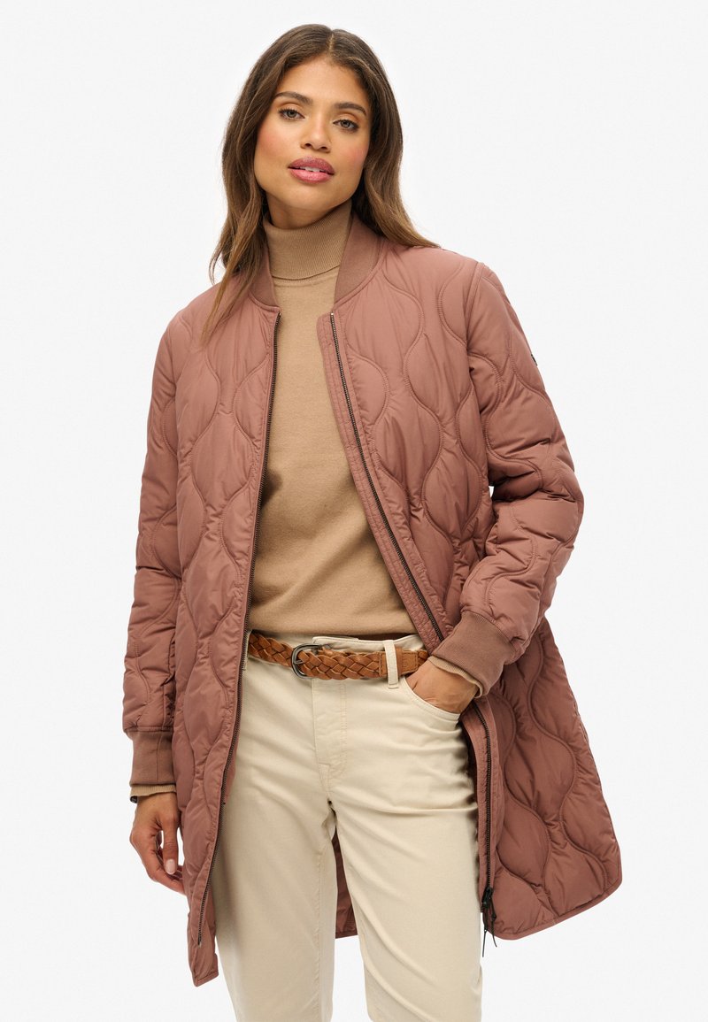Femme portant une veste bomber matelassée mauve sur un pull à col roulé beige, associée à un pantalon beige clair et une ceinture tressée marron.