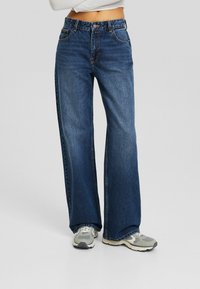 Wide-leg jeans in medium blue denim, featuring contrasting stitching, front pockets, and a button closure. Paired with gray sneakers.