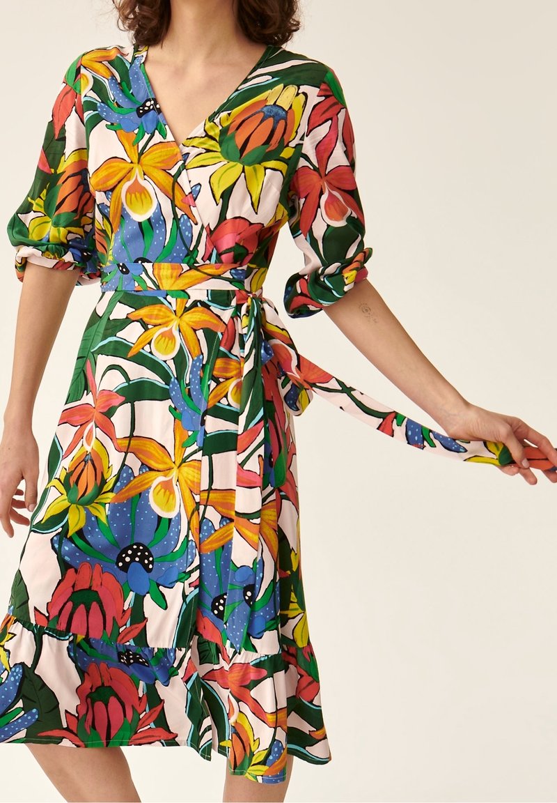 Femme portant une robe portefeuille fleurie colorée à manches trois-quarts nouée à la taille, tenant une extrémité de la ceinture.