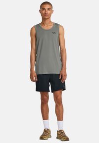 Graues Tanktop mit dezentem Logo, kombiniert mit schwarzen Shorts. Tragen mit weißen Socken und braunen Sportschuhen mit bunten Schnürsenkeln.