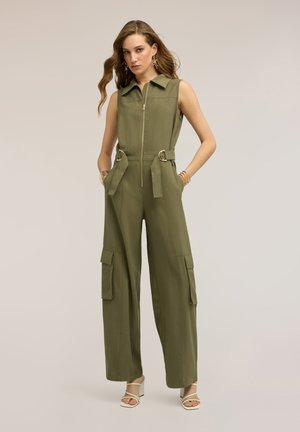 Kvinde stående med hænderne i lommerne iført ærmeløs olivengrøn jumpsuit med cargolommer og hvide høje sandaler.