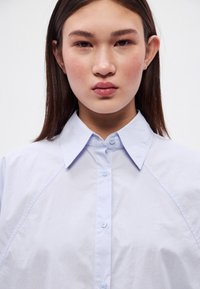 Ανοιχτό μπλε πουκάμισο με κουμπιά και δομημένο γιακά, με ομαλή υφή υφάσματος και εφαρμοστή γραμμή, τονισμένο από εμφανή κουμπιά.