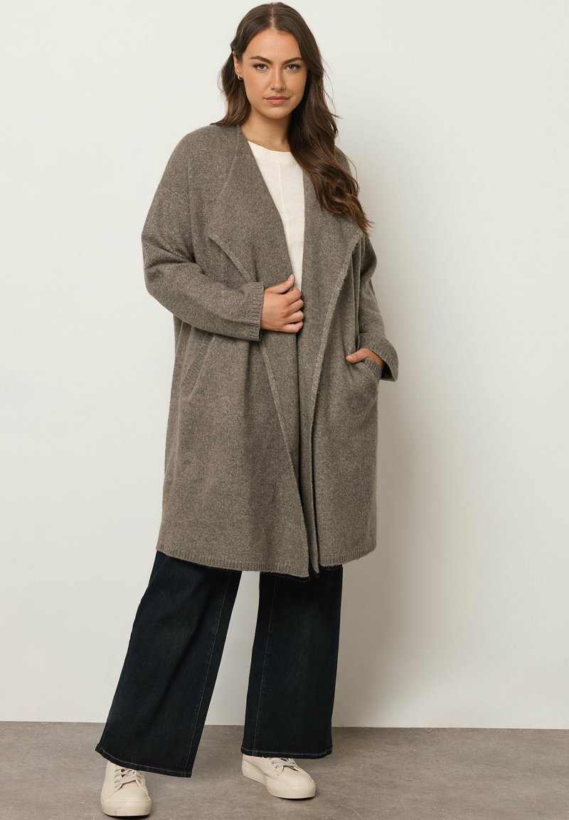 Cardigan in maglia oversize grigio chiaro, con apertura frontale, tasche e maniche lunghe, abbinato a jeans larghi scuri e sneakers crema.