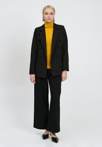 Zwarte dubbelzijdige blazer over een felgele coltrui, gecombineerd met brede zwarte broekspijpen en puntige zwarte instappers.