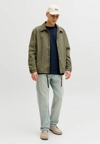 Lätt olivgrön jacka med avslappnad passform, elastiska muddar och dragsko i nederkant, kombinerad med en marinblå skjorta, ljusa jeans och beige skor.