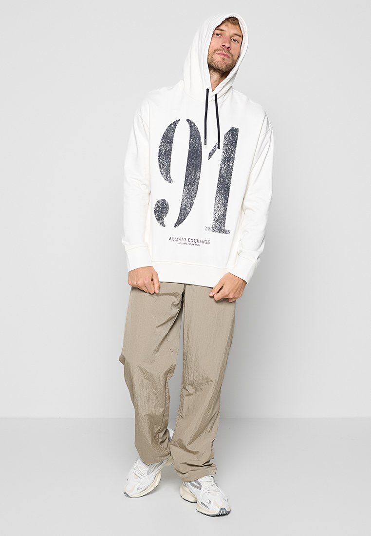 Sudadera blanca con capucha y un gran gráfico negro "91", combinada con pantalones de corte relajado de color marrón claro y zapatillas blancas. Diseño simple.