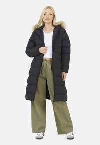 Long manteau en duvet noir avec une capuche bordée de fourrure, caractérisé par un design à cloisons, associé à un pantalon large olive et des baskets.