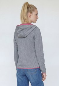 Femme à queue de cheval blonde portant un sweat à capuche gris avec des bordures roses et un jean bleu, vue de profil sur un fond uni clair.
