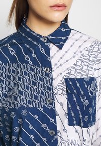 Chemise à motifs bleu marine et blanc avec un design à boutons, des tissus contrastés et une poche poitrine ornée de motifs géométriques et de cordes.