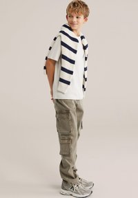 Bijela majica kratkih rukava uparena s maslinasto zelenim cargo hlačama više džepova i krem kardiganom s tamnoplave pruge prebačenim preko ramena.