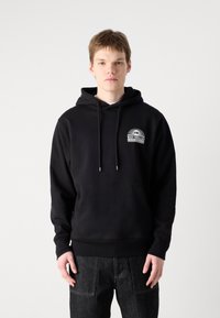 Sudadera con capucha negra y un bolsillo delantero, con un logo blanco en el lado izquierdo. El material parece suave y de textura lisa.