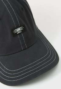 Mørkegrå baseballcap laget av stoff, med hvite sømmer og en svart logo-patch med flammedesign foran.