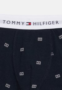 Σκούρα μπλε μεταξωτά μπόξερ με μικρά λευκά ορθογώνια λογότυπα και λευκή ζώνη μέσης με το κείμενο "Tommy Hilfiger" και το λογότυπο στο κέντρο.