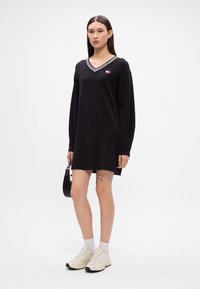 Μαύρο φόρεμα μακρυμάνικο με V-λαιμόκοψη, με λευκές ρίγες. Το μοντέλο φοράει ανοιχτόχρωμα αθλητικά παπούτσια και κρατάει μια μικρή μαύρη τσάντα. Αδιόρατο τατουάζ ορατό.