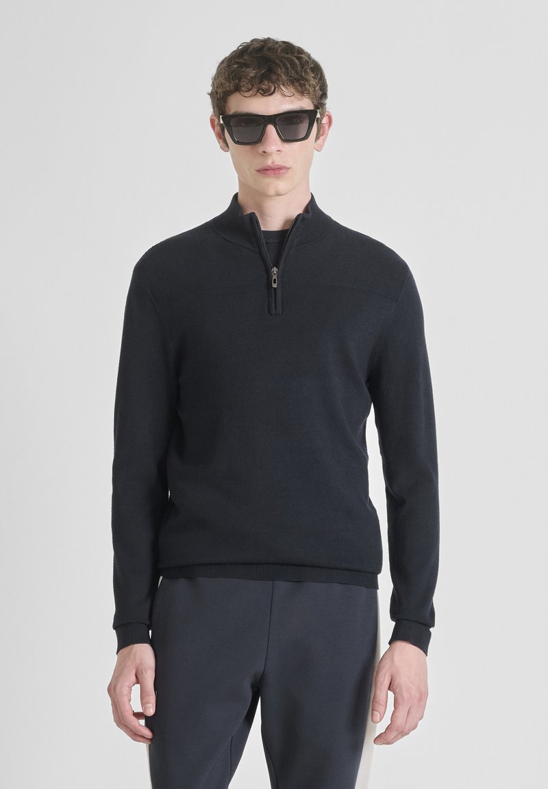 Joven vestido con un suéter negro de media cremallera, pantalones oscuros y grandes gafas de sol negras, de pie frente a un fondo blanco liso.