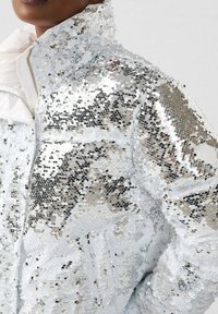 Close-up of person wearing a silver sequin jacket with a high collar and a white inner lining.