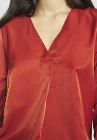 Blouse rouge à encolure en V avec une légère brillance. Texture douce et lisse, présentant un design légèrement drapé et des détails minimalistes.