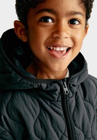 Bambino sorridente con capelli ricci, che indossa una giacca imbottita scura con zip frontale e cappuccio, su uno sfondo neutro.