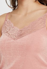 Camisole rose avec des bretelles fines, des détails en dentelle au niveau du col et un tissu doux, légèrement texturé.