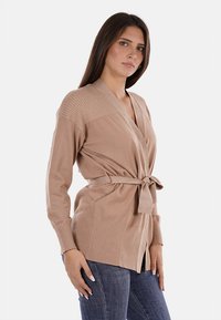 Cardigan beige in maglia con frontale avvolgente e cintura, caratterizzato da una texture a coste sulle spalle e sulle maniche, abbinato a jeans in denim.