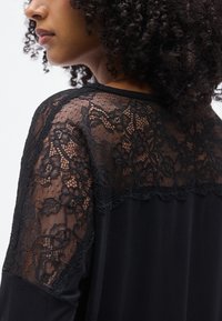 Μαύρο τοπ με διαφανή δαντελένιο επικάλυμμα στους ώμους, που παρουσιάζει λουλουδάτα σχέδια και υφασματικές λεπτομέρειες πάνω σε μια μονόχρωμη βάση υφάσματος.