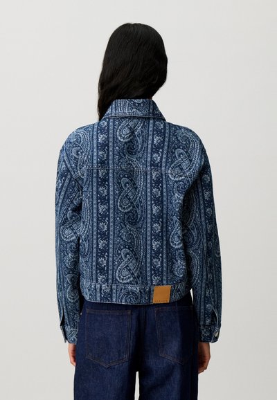 Persona con capelli lunghi e scuri indossa una giacca blu fantasia e pantaloni blu scuro, in piedi di spalle su uno sfondo chiaro e semplice.