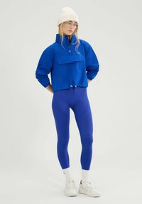 Blauwe cropped pullover met een voorkeurszak en een taille met trekkoord, gecombineerd met blauwe legging met textuurpatronen en witte sneakers.