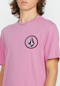 Pink cotton t-shirt featuring a circular black logo with a stylized geometric design. Short sleeves and crew neck. Smooth texture.