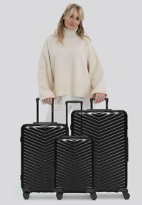 Femme portant un pull à col roulé beige se tenant derrière trois valises rigides noires de différentes tailles.