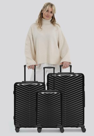 Femme portant un pull à col roulé beige se tenant derrière trois valises rigides noires de différentes tailles.