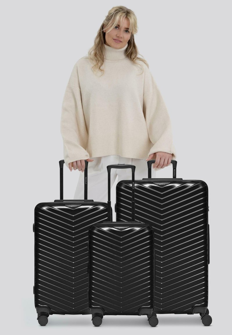 Vrouw in een beige coltrui staat achter drie zwarte harde koffers in verschillende maten.