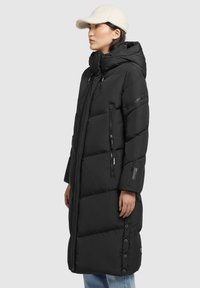Cappotto lungo imbottito nero con cappuccio, tasche con cerniera e bottoni automatici. Presenta una texture liscia e una vestibilità comoda. Indossato con un berretto beige.