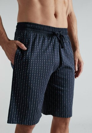 Man wearing navy blue patterned drawstring shorts with hands near pockets against plain gray background.