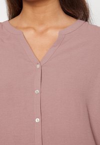 Blusa rosa chiaro con scollo a V, dotata di una patta con bottoni, cuciture sottili e una texture di tessuto morbida e liscia.