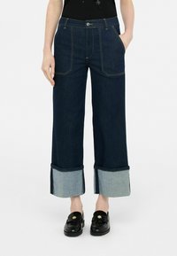 Pantalones de mezclilla de pierna ancha en azul oscuro con un dobladillo azul claro. Presenta bolsillos delanteros y costuras en contraste. Combinados con mocasines negros.