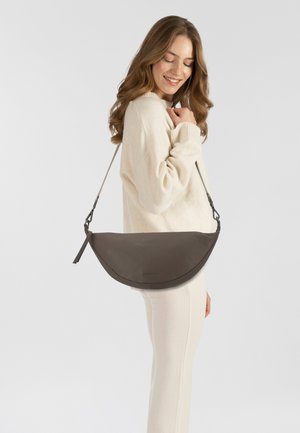 Sac à bandoulière en cuir marron avec un design incurvé, une bandoulière ajustable et un détail de frange. Porté en travers du corps, mettant en avant une texture lisse.