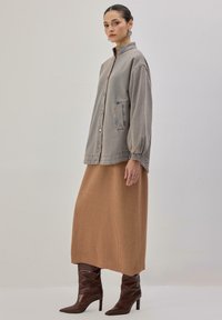 Siva oversized jakna iz jeansa z gumbi, elastičnimi rokavi in prsnim žepom. Model nosi rjavo rebrasto maxi krilo in rjave konične škornje.